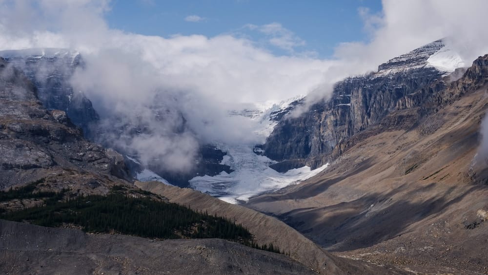 El glaciar Athabasca desciende entre paredes rocosas de montaña en Columbia Icefield, Alberta, Canadá, con sectores de bosque más abajo y nubes bajas cruzando el hielo y las cumbres.