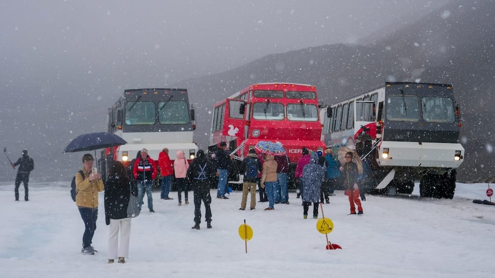 Visitantes con diferentes tipos de abrigo durante una tormenta de nieve en Columbia Icefield — Glaciar Athabasca, Alberta Una multitud de visitantes en el glaciar Athabasca durante una fuerte nevada vistiendo una gran variedad de ropa de abrigo, desde camperas térmicas hasta paraguas sobre ropa de calle, con tres vehículos Ice Explorer estacionados detrás, Alberta, Canadá.