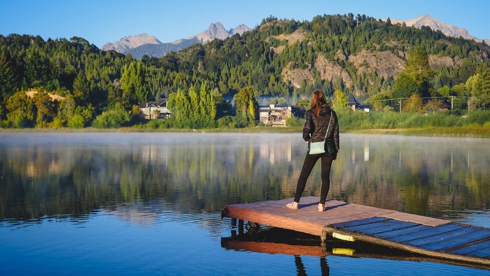 Audrey Bergner está sobre un muelle de madera junto a aguas alpinas calmas en Bariloche, con colinas boscosas, picos y casas costeras reflejadas en el lago, Río Negro, Argentina.