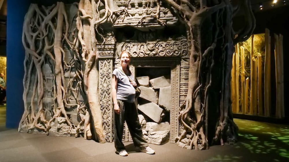 Audrey Bergner en la exhibición de Angkor Wat (ahora cerrada), Royal Alberta Museum, Edmonton, Alberta, Canadá Audrey Bergner posa sonriente frente a la fachada de una réplica de un templo de Angkor Wat entrelazada con raíces de árboles, parte de una exhibición especial que alguna vez se mostró en el Royal Alberta Museum en Edmonton, Alberta, Canadá.
