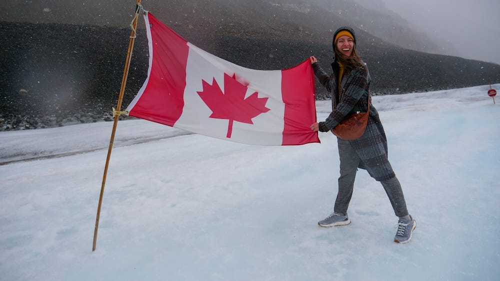 Audrey Bergner sostiene una gran bandera canadiense mientras está parada sobre el glaciar Athabasca en Columbia Icefield, Alberta, Canadá, con nieve cayendo y laderas oscuras perdiéndose en la niebla.