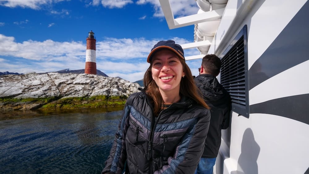 Audrey Bergner pasando frente al Faro Les Éclaireurs en el Canal Beagle, Ushuaia, Argentina Audrey Bergner sonriendo en la cubierta de un barco con el faro a rayas rojas y blancas Les Éclaireurs y un islote rocoso cubierto de musgo detrás de ella, navegando el Canal Beagle cerca de Ushuaia, Tierra del Fuego, Patagonia argentina.