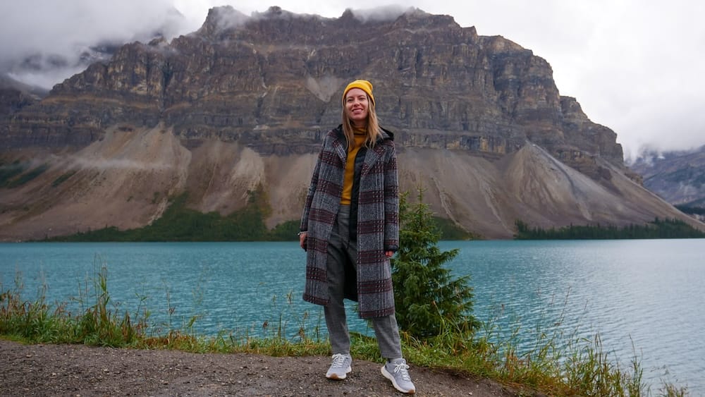 Audrey Bergner stands on the gravel shoreline of Bow Lake along the Icefields Parkway in Banff National Park, Alberta, with a small evergreen and the turquoise water behind her beneath a cloud-covered peak.