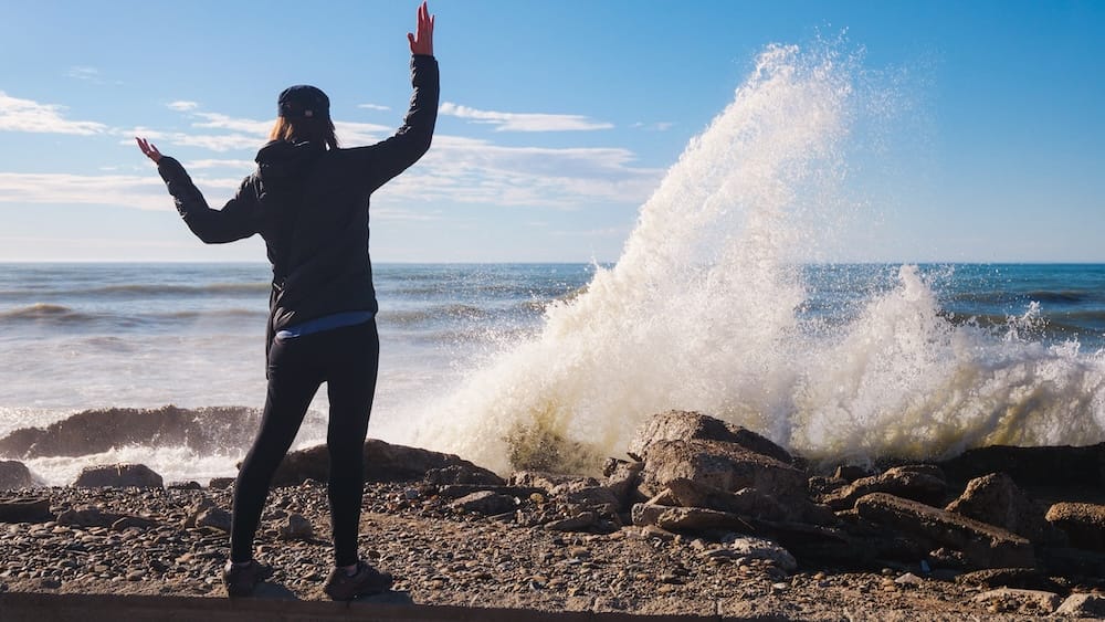 Audrey Bergner standing on a rocky shoreline in Comodoro Rivadavia, Chubut, Argentina as powerful Atlantic waves crash against the coast, capturing the wind-swept energy and raw coastal landscape of Patagonia’s rugged seaside.
