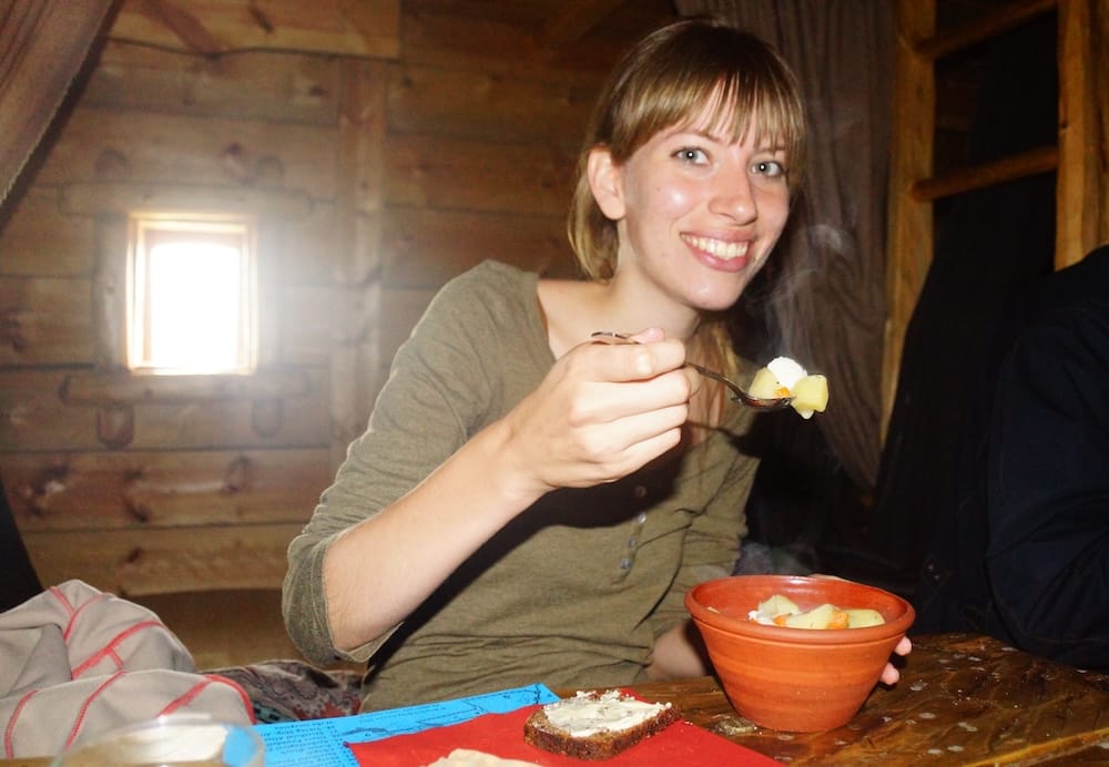 Audrey Bergner smiling while eating a hearty Finnish meal from a bowl beside a slice of rye bread inside a rustic wooden cabin, capturing a warm and personal food moment during our travels in Finland, Audrey Bergner.
