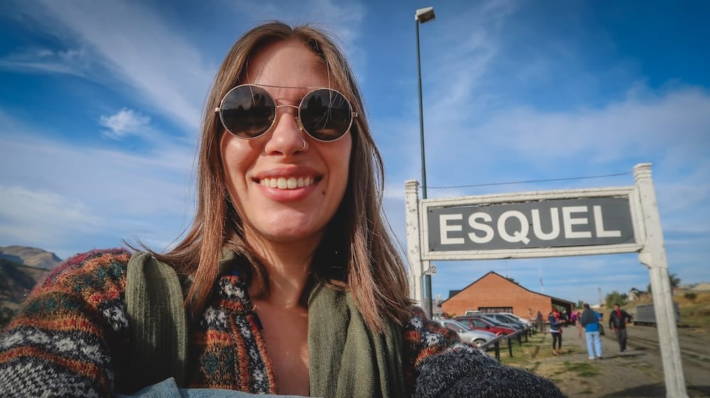 Audrey Bergner at Esquel Railway Station and La Trochita Terminal Logistics in Chubut Patagonia Audrey Bergner at Esquel railway station beside the La Trochita terminal sign in Chubut, showing how Patagonia travel times depend on station logistics, ticket windows and local schedules as much as distance.