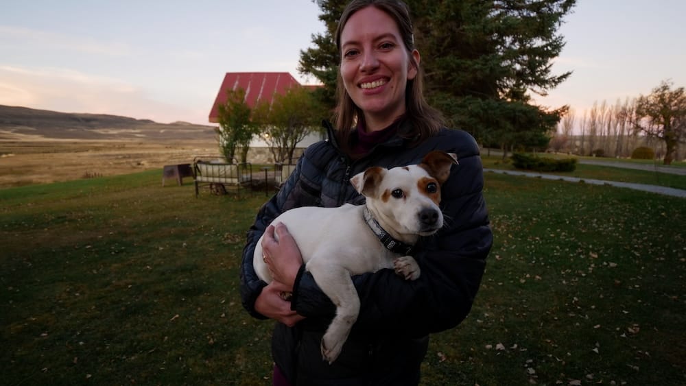 Audrey Bergner smiles while holding the lodge's resident dog at golden hour at Estancia Tecka near Esquel, Chubut, Argentine Patagonia, with the ranch's red-roofed outbuilding and open grassland behind her.