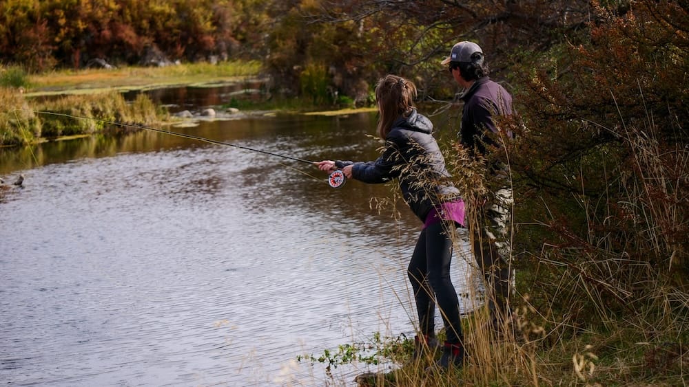 Audrey Bergner casts a fly rod on the Río Corcovado with her guide beside her at Estancia Tecka near Esquel, Chubut, Argentine Patagonia, autumn foliage lining the riverbank behind them.