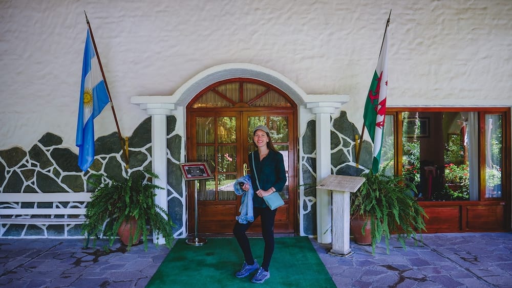 Audrey Bergner standing outside a historic Welsh tea house in Gaiman, Argentina, with Argentine and Welsh flags highlighting the cultural heritage of Patagonia shaped by settlement patterns within the arid steppe environment.
