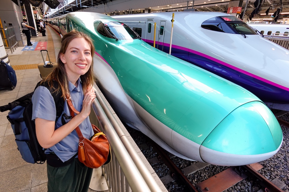 Audrey Bergner Beside the Hokkaido Shinkansen Hayabusa Bullet Train Before Travelling from Tokyo to Hakodate, Japan Audrey Bergner smiling beside a turquoise-green Hokkaido Shinkansen bullet train on a Japanese railway platform, with a white and purple Shinkansen on the adjacent track and passengers waiting in the background.