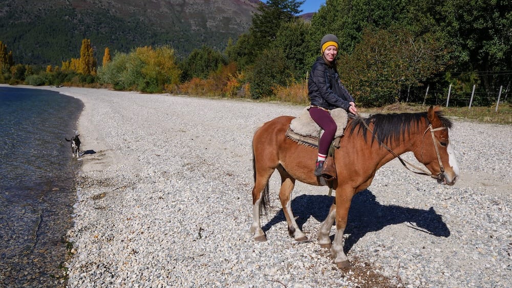 Audrey Bergner rides a horse along the pebbled shore of Lago Gutiérrez near Bariloche, Río Negro, Patagonia, Argentina, with autumn trees, mountain slopes and a dog following beside the lakeside trail.