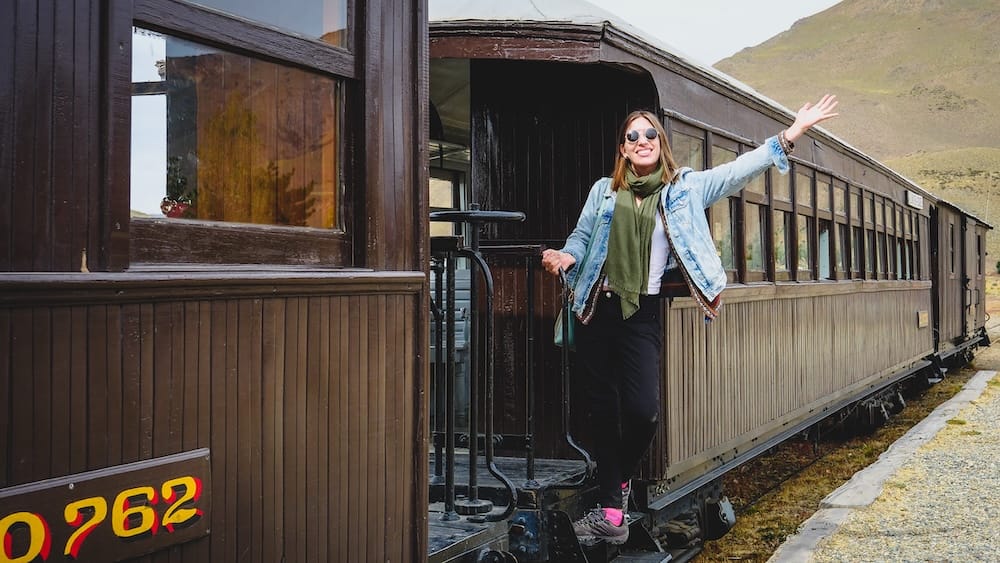 Audrey Bergner aboard La Trochita at the Old Patagonian Express station in Esquel, Patagonia, Argentina Audrey Bergner stands on the steps of a wooden La Trochita carriage at the historic Old Patagonian Express station in Esquel, with dry hills rising behind her in Chubut, Patagonia, Argentina.
