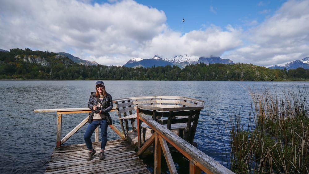 Audrey Bergner está sobre una plataforma rústica de madera junto al lago Nahuel Huapi, con costa boscosa, juncos y picos nevados cerca de Bariloche, Río Negro, Patagonia argentina.