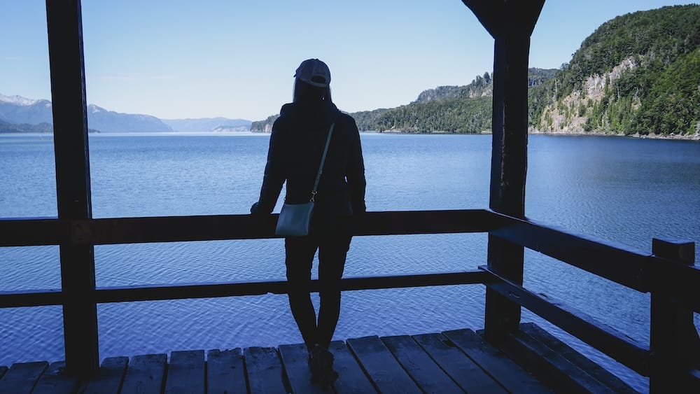 Lake viewpoint stop along the Seven Lakes Route, Neuquén/Río Negro, Patagonia, Argentina Audrey Bergner stands in silhouette against a still Andean lake, leaning on a wooden railing at a viewpoint stop along the Seven Lakes Route in Neuquén/Río Negro, Patagonia, Argentina.