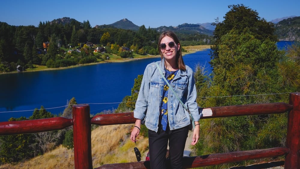Audrey Bergner at a Viewpoint on the Walk Up to Llao Llao Hotel, Bariloche, Río Negro, Patagonia Audrey Bergner smiles beside a wooden railing overlooking a blue lake, forest, and distant mountains at a viewpoint along the long hilly entrance road up to Llao Llao Hotel in Bariloche, Río Negro, Argentine Patagonia, Argentina.
