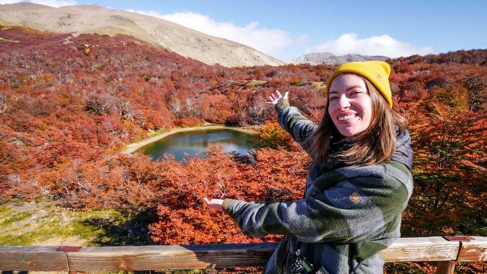 Audrey Bergner sonríe y señala Laguna Verde, rodeada de intenso follaje otoñal rojo y naranja cerca del Lago Gutiérrez en Bariloche, Río Negro, Patagonia argentina.
