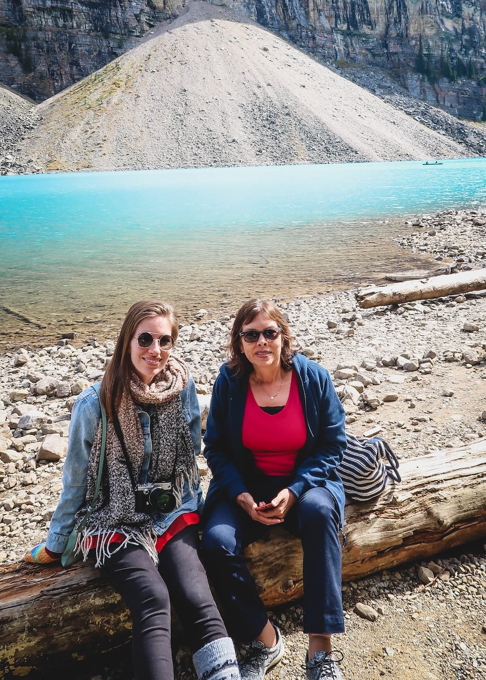 Audrey Bergner sits with her mother on a driftwood log along the quiet shoreline of Moraine Lake in Banff National Park, Alberta, Canada, with a distant canoe visible on the turquoise water.