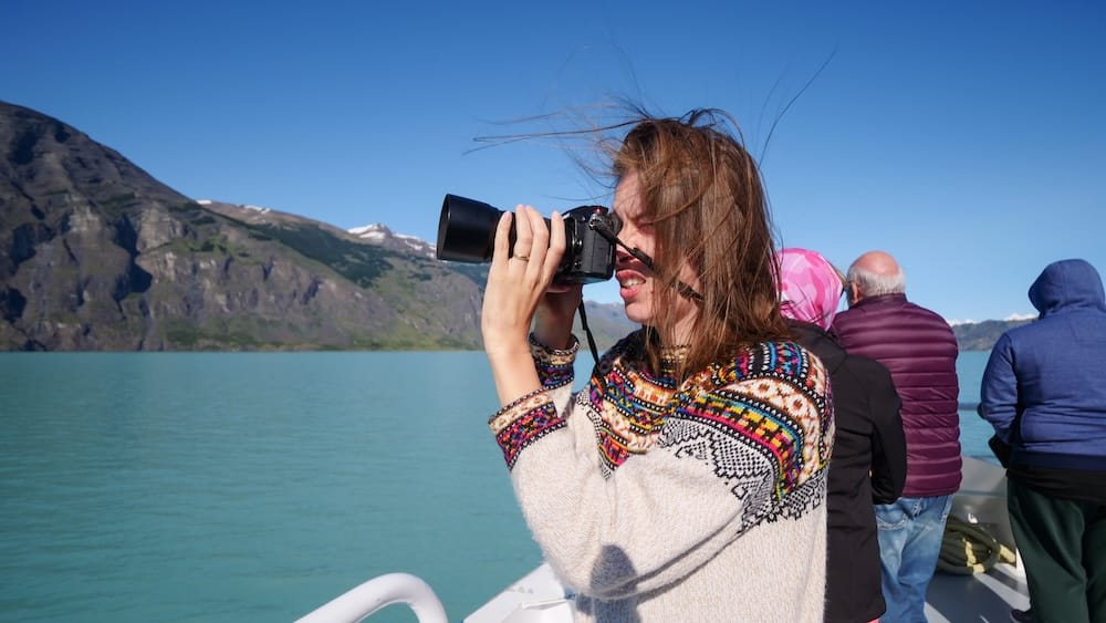 Audrey Bergner photographing the glacial waters of Perito Moreno in El Calafate, Argentina, with strong Patagonian winds blowing her hair, illustrating the powerful environmental forces shaping the Andean landscape.