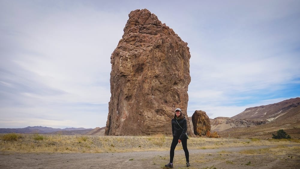 Audrey Bergner stands beneath the towering volcanic monolith of Piedra Parada in Chubut, Patagonia, Argentina, surrounded by barren steppe, eroded hills and a vast open sky.