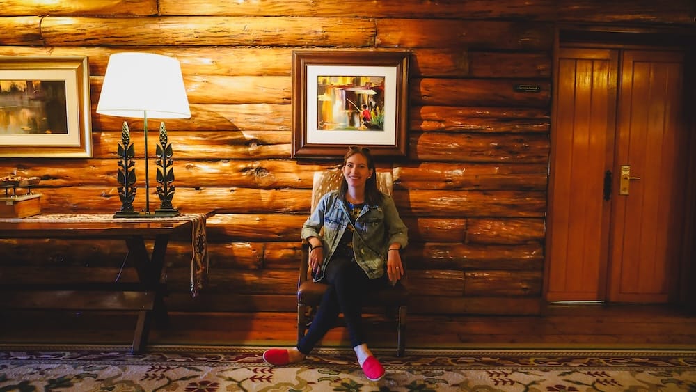 Audrey Bergner in the Historic Log-Cabin Lobby of Llao Llao Hotel, Bariloche, Río Negro, Argentine Patagonia Audrey Bergner sits in a wooden armchair against a log-cabin wall in the Llao Llao Hotel's lobby in Bariloche, Río Negro, Argentine Patagonia, wearing red slippers beside a carved wooden table lamp and framed local artwork.