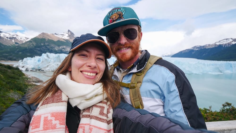 Audrey Bergner y Samuel Jeffery sacándose una selfie frente a las macizas e icónicas formaciones de hielo azul del Glaciar Perito Moreno en Santa Cruz, Patagonia, Argentina. Están sonriendo y vestidos con ropa técnica de invierno, incluyendo bufandas y gorras.