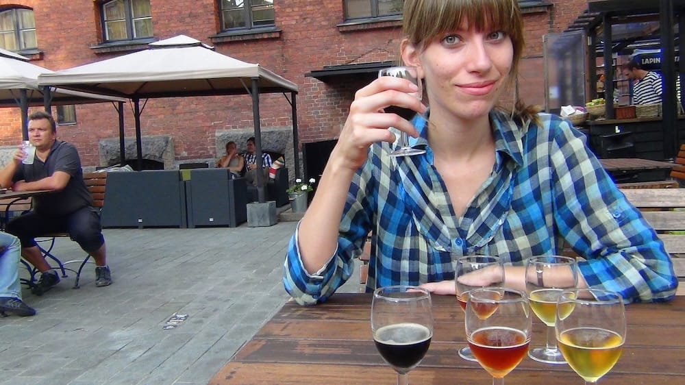 Audrey Bergner holds a dark Finnish craft beer while a tasting flight of pale, amber and dark beers sits on an outdoor terrace table in Helsinki, documenting our informal Finnish beer experience, Audrey Bergner.
