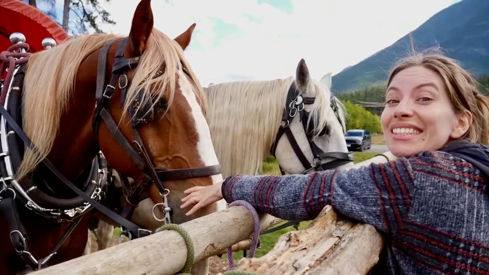 Audrey Bergner con caballos de carro, Banff, Alberta, Canadá Audrey Bergner sonríe mientras acaricia a uno de los dos caballos de tiro atados a un carro techado en Banff, Alberta, Canadá, con montañas visibles de fondo.