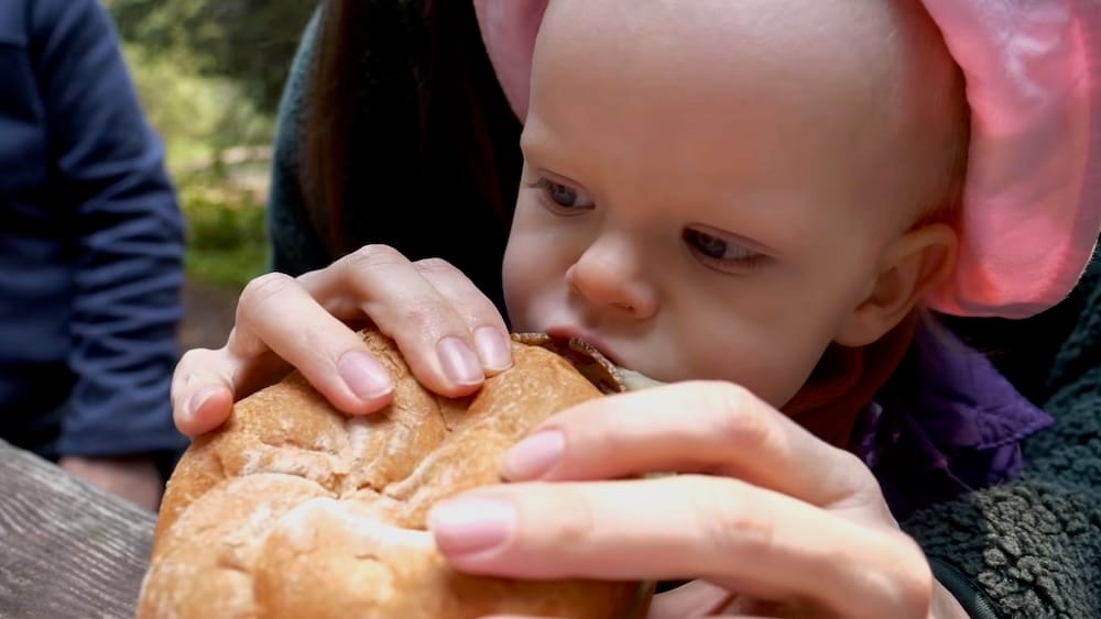 Aurelia Jeffery eating a large roast beef sandwich during a family outing near Lake Louise in Banff National Park, Alberta, Canada, with an adult helping hold the sandwich in the foreground.