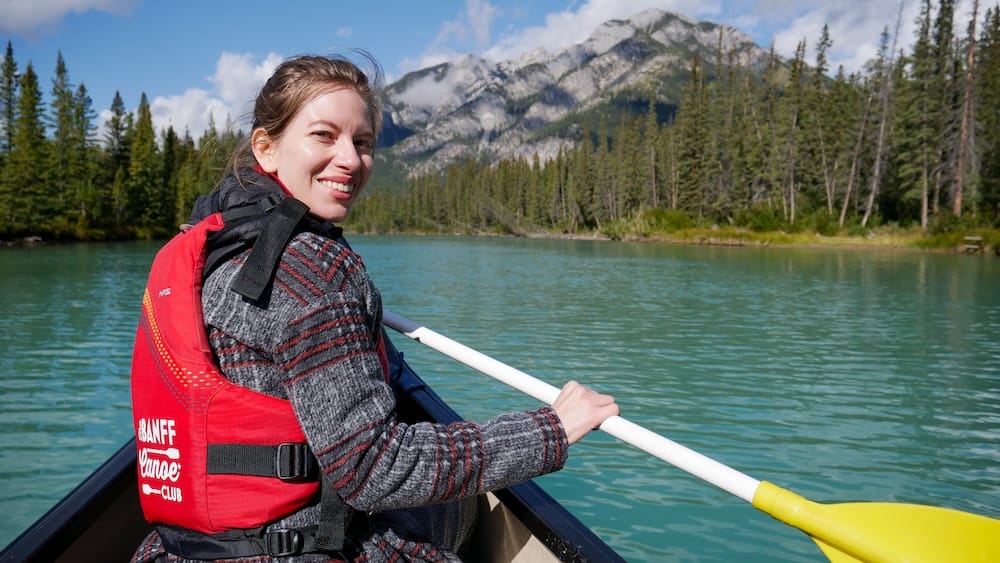 Audrey Bergner Paddling with Banff Canoe Club on the Bow River in Banff, Alberta, Canada Audrey Bergner paddling a canoe on the Bow River with Banff Canoe Club in Banff, Alberta, Canada, wearing a red life jacket with turquoise water, forest and Rocky Mountain peaks behind her.