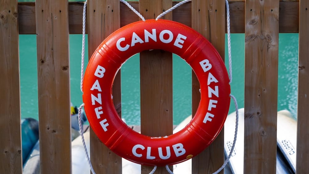 Banff Canoe Club junto al río Bow en Banff, Alberta, Canadá, con salvavidas rojo y equipo para alquiler de canoas Salvavidas circular rojo del Banff Canoe Club colocado sobre una cerca de madera junto al río Bow de aguas turquesas en Banff, Alberta, Canadá, con cascos de canoas y equipo del muelle visibles detrás.