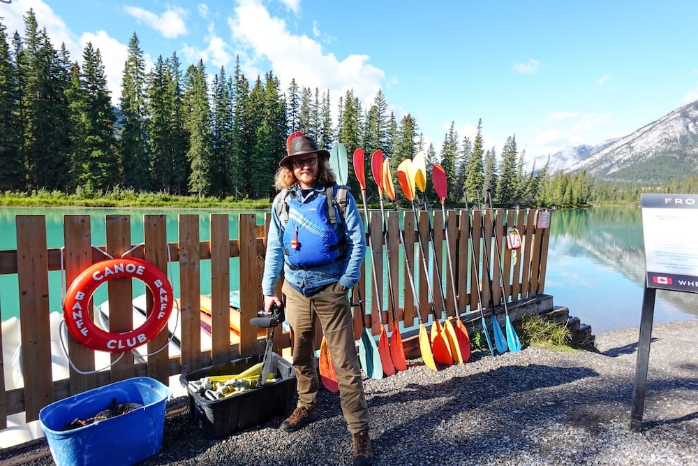 Samuel Jeffery listo para remar en Banff Canoe Club, junto al río Bow en Banff, Alberta, Canadá Samuel Jeffery en el Banff Canoe Club junto al río Bow en Banff, Alberta, Canadá, con un chaleco salvavidas azul, remos de colores, canoas de alquiler y paisaje de montaña detrás.