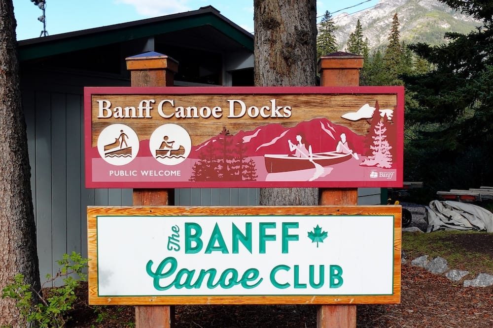 Los muelles y el cartel del Banff Canoe Club en el punto de encuentro compartido para el Big Canoe Tour en Banff, Alberta, Canadá, con una montaña visible detrás de los árboles.
