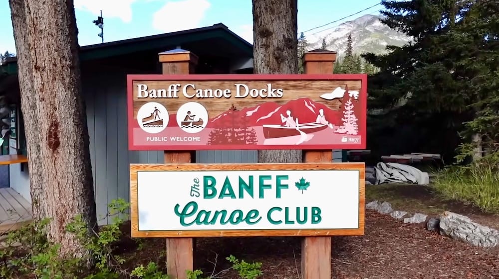 El cartel del Banff Canoe Club y los muelles de canoas de Banff en el punto de encuentro del tour en Banff, Alberta, Canadá, con una montaña asomando entre los árboles detrás de la cabaña de madera.