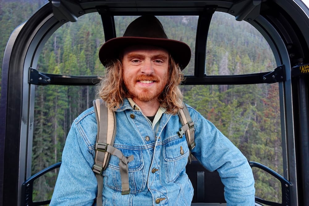 Samuel Jeffery riding the Banff Gondola in Banff National Park, Alberta, Canada, with forest views behind him Samuel Jeffery rides inside a Banff Gondola cabin in Banff National Park, Alberta, Canada, with evergreen forest and Rocky Mountain scenery visible through the curved windows behind him.