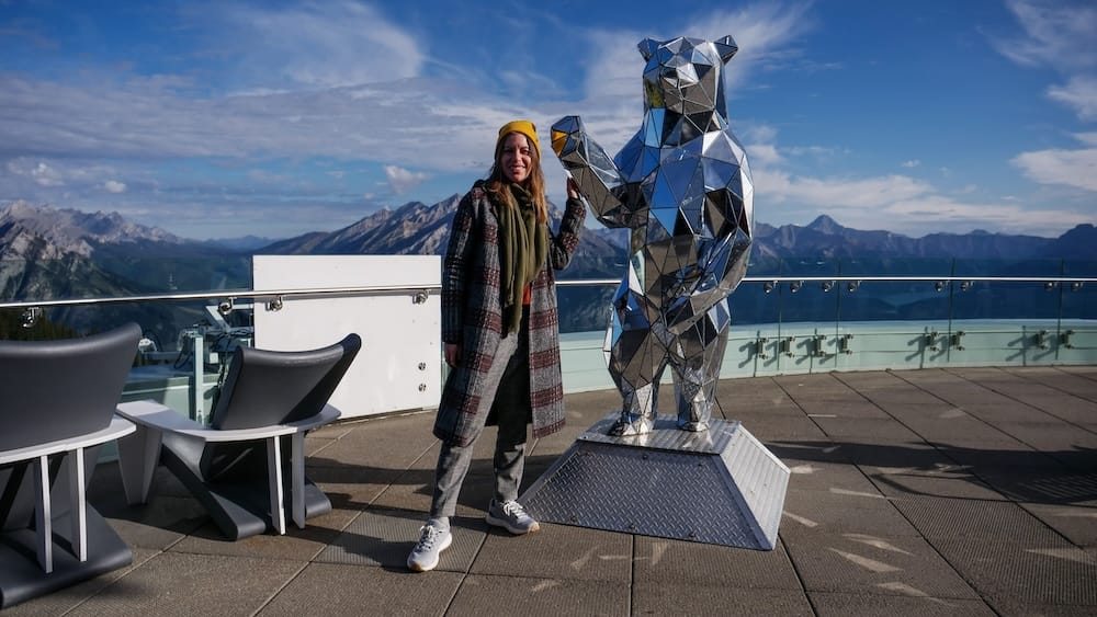 Audrey Bergner on the Banff Gondola rooftop observation deck, Sulphur Mountain, Banff, Alberta, Canada Audrey Bergner stands beside a mirrored bear sculpture on the Banff Gondola's rooftop observation deck, with clear blue sky and layered mountain ranges visible on Sulphur Mountain, Alberta, Canada.