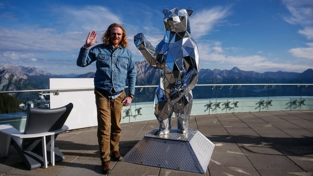 Samuel Jeffery posing beside a mirrored bear sculpture after riding the Banff Gondola on Sulphur Mountain in Alberta, Canada, with panoramic Canadian Rockies peaks behind him.