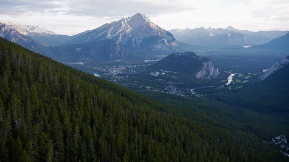 Scenic view from the Banff Gondola ascent on Sulphur Mountain, looking across Banff, Alberta, Canada, with forested slopes, the townsite, winding river and rugged Rocky Mountain peaks beyond.