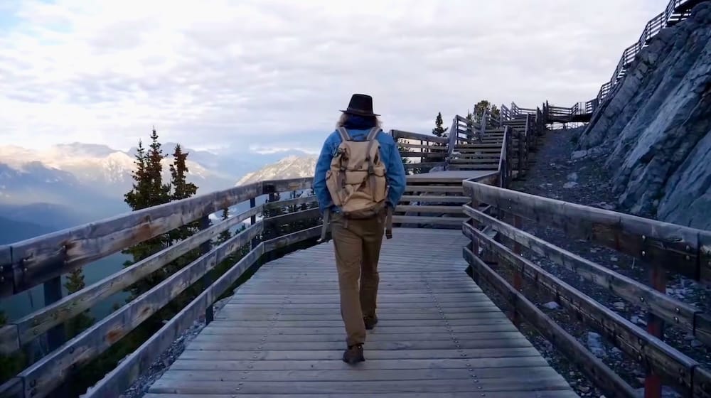 Samuel Jeffery walking along the wooden summit boardwalk on Sulphur Mountain at the Banff Gondola in Banff, Alberta, Canada, with mountain views opening beyond the railing and rocky terrain beside the path.