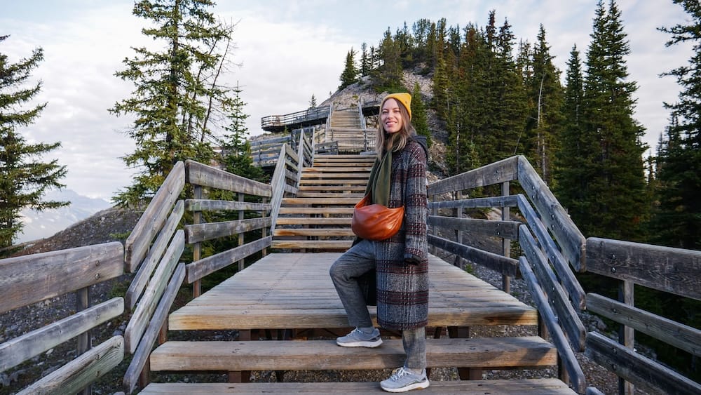 Audrey Bergner en las escaleras de la pasarela de Sulphur Mountain sobre Banff, Alberta, Canadá, cerca de la cumbre de la Banff Gondola Audrey Bergner sobre la pasarela de madera de Sulphur Mountain, arriba de Banff, Alberta, Canadá, con amplios tramos de escaleras, barandas de madera, bosque de coníferas y terreno rocoso elevándose detrás.