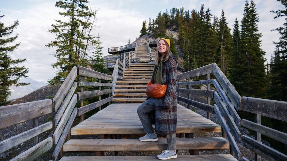 Audrey Bergner standing on the wooden summit boardwalk and stairs at the Banff Gondola on Sulphur Mountain in Banff, Alberta, Canada, surrounded by evergreen trees and rocky mountain terrain.