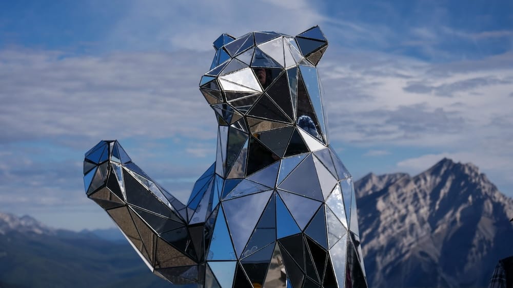 Mirrored geometric bear sculpture waving at the Banff Gondola summit on Sulphur Mountain in Banff, Alberta, Canada, with rugged Rocky Mountain peaks and blue sky in the background.