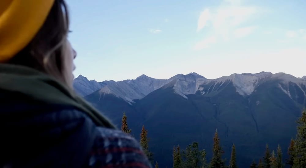 Audrey Bergner disfrutando las vistas de montaña desde Sulphur Mountain sobre Banff, Alberta, Canadá, cerca de la cumbre de la Banff Gondola Audrey Bergner contemplando escarpadas cordilleras desde la cumbre de la Banff Gondola en Sulphur Mountain sobre Banff, Alberta, Canadá, con laderas boscosas oscuras y capas de las Montañas Rocosas extendiéndose por el horizonte.