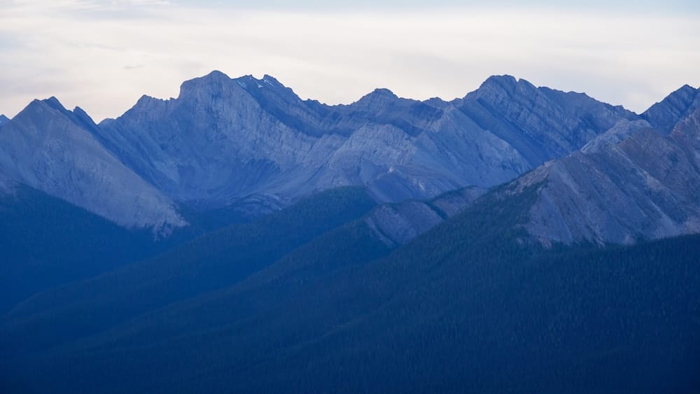 Layered Rocky Mountain peaks viewed from Sulphur Mountain near the Banff Gondola in Banff, Alberta, Canada, with dark forested slopes below and soft cloud cover creating cool, moody mountain light.