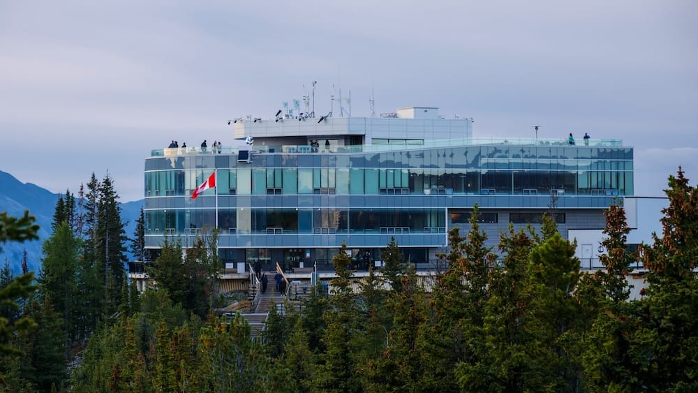 Banff Gondola summit building on Sulphur Mountain in Banff, Alberta, Canada, rising above evergreen forest with glass viewing decks, rooftop visitors and mountain scenery in the background.
