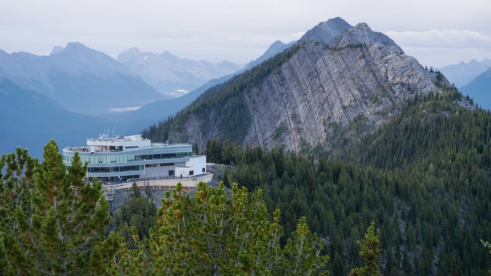 Edificio de la cumbre de la Banff Gondola en Sulphur Mountain sobre Banff, Alberta, Canadá, con vistas de las Montañas Rocosas Edificio de la cumbre de la Banff Gondola en Sulphur Mountain sobre Banff, Alberta, Canadá, rodeado de bosque de coníferas, una empinada cresta rocosa y capas de las Montañas Rocosas canadienses perdiéndose en la distancia.