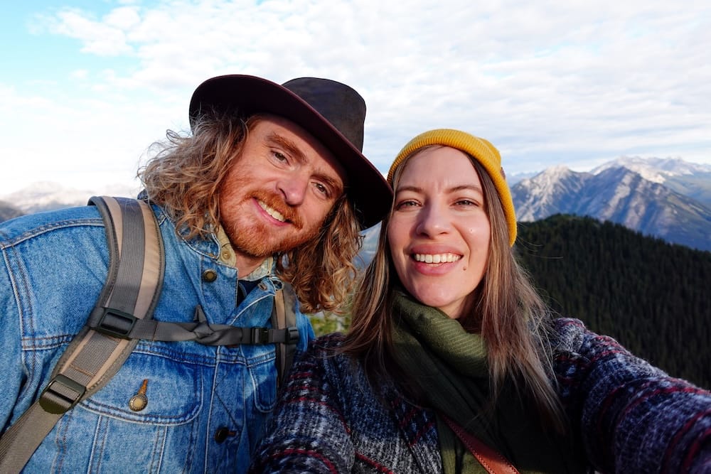 Samuel Jeffery and Audrey Bergner taking a selfie at the Banff Gondola summit on Sulphur Mountain in Banff, Alberta, Canada, with a forested ridge and Rocky Mountain peaks visible behind them.
