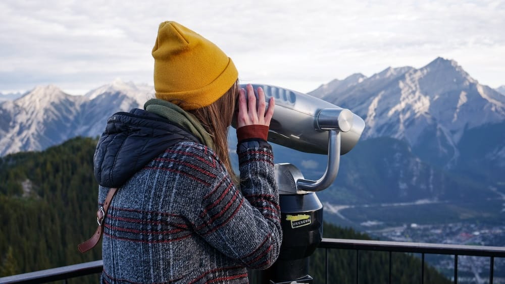 Audrey Bergner looking through a mounted viewing scope at the Banff Gondola summit on Sulphur Mountain in Banff, Alberta, Canada, with rugged Rocky Mountain peaks and the valley below.
