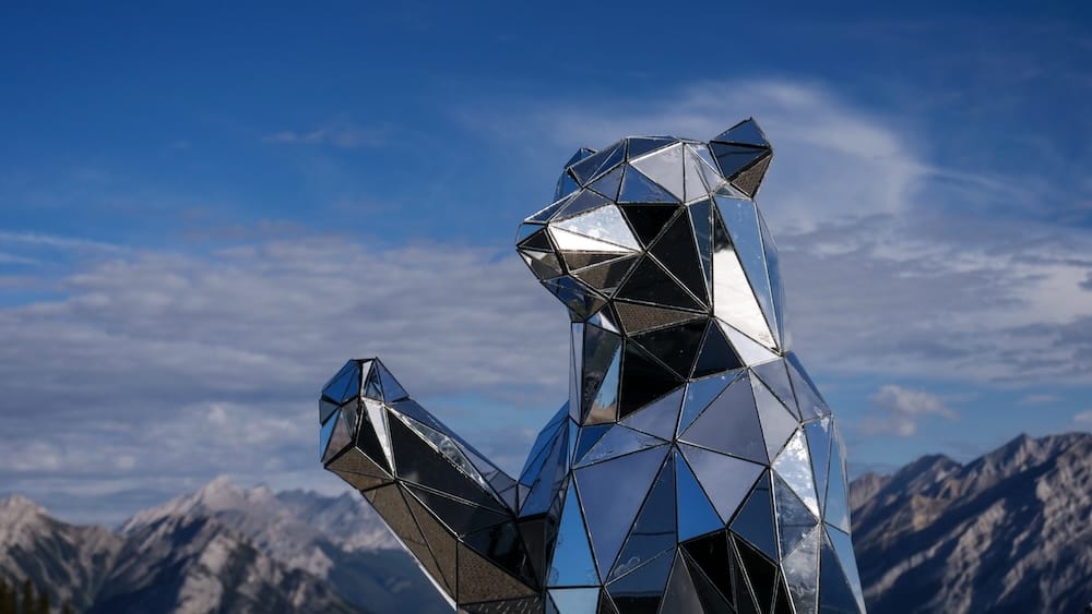 Reflective geometric bear sculpture raising one paw at the Banff Gondola summit on Sulphur Mountain in Banff, Alberta, Canada, with rugged Canadian Rockies and blue sky behind it.