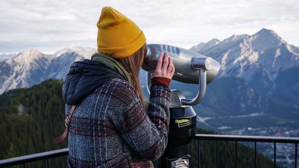 Audrey Bergner looking through a viewing scope at the Banff Gondola summit on Sulphur Mountain in Banff, Alberta, Canada, with rugged Rocky Mountain peaks and part of Banff visible below.