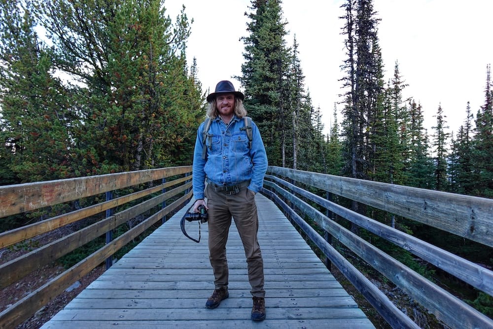 Samuel Jeffery standing on the wooden boardwalk at the Banff Gondola summit on Sulphur Mountain in Banff, Alberta, Canada, holding a camera with evergreen forest surrounding the walkway.